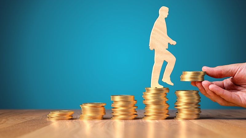 Wooden cutout of a human walks up a pile of gold coins